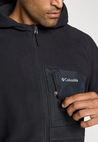 Man wearing black Columbia fleece hoodie, holding and partially unzipping small chest pocket zipper.