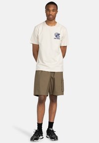 Beige Baumwoll-T-Shirt mit navyblauem Grafikdesign, kombiniert mit khakifarbenen Cargo-Shorts mit Seitentaschen. Schwarze Sportschuhe runden das Outfit ab.