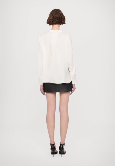 Femme aux cheveux courts et bruns, portant un chemisier blanc à manches longues, une mini-jupe en cuir noire et des sandales noires à talons hauts, vue de dos.