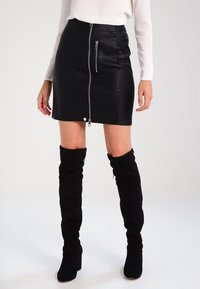 Mustat tekonahkainen minihame, jossa on edessä vetoketjusuljenta, ja metallinen vetoketju yksityiskohtana. Yhdistetty polvipitkiin mustiin saappaisiin.