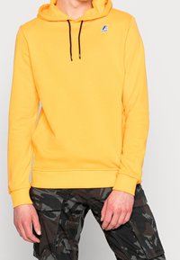 Żółta bluza z kapturem i sznurkiem, z logo na klatce piersiowej. W zestawie z czarnymi spodniami cargo w kamuflażu, ukazującymi kieszenie i teksturę materiału.