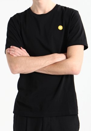 Czarna bawełniana koszulka z okrągłym dekoltem i krótkimi rękawami; na lewej piersi znajduje się małe żółte logo. Płaska, gładka tekstura.