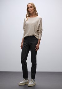 Beżowy sweter dzianinowy o szerokim dekolcie, zestawiony z czarnymi, dopasowanymi spodniami. Model nosi jasne trampki. Tło jest jednolicie białe.