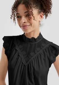 Blusa nera con ricami floreali su un colletto alto, maniche corte con rouches e tessuto morbido e fluido con dettagli plissettati.