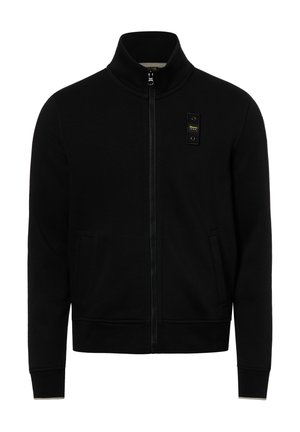 Schwarze Jacke mit Reißverschluss, hohem Kragen, zwei Fronttaschen und einem kleinen rechteckigen Aufnäher mit gelbem Text auf der linken Brust.