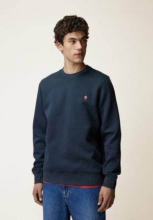 Giovane uomo con capelli ricci che indossa una felpa blu navy sopra una camicia rossa e jeans blu, che guarda verso sinistra con uno sfondo semplice.