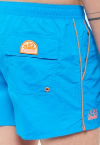 Pantaloni da bagno blu con vita elastica, dettagli arancioni e una tasca posteriore con una toppa di marchio arancione circolare.