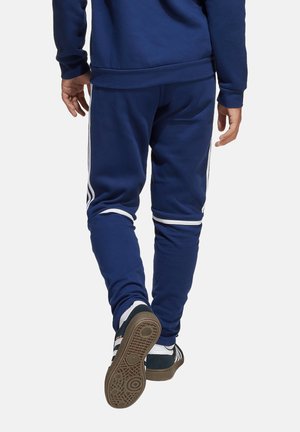 Pantalons de jogging bleu marine en tissu doux, avec des rayures blanches sur les côtés et un design fuselé. Portés avec des baskets noires à semelles texturées.