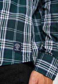 Zeleno-námořnická modro-bílá kostkovaná košile s manžetou a postranním lemem, na zápěstí osoby je logo Tom Tailor, viditelné jsou také černé kalhoty.