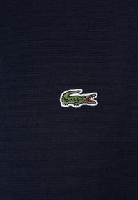 Tumma laivastonsininen kangas, jolla on pieni kirjailtu vihreä krokotiililogo punaisilla korostuksilla, asetettuna teksturoidulle taustalle.