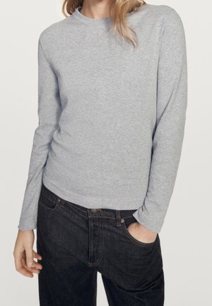 Femme portant une chemise gris clair à manches longues et un jean noir, une main dans la poche, sur un fond uni.