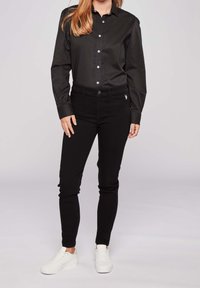 Černá košile na knoflíky s bílými knoflíky, spárovaná se slim-fit černými džíny a bílými teniskami. Materiál vypadá hladce s minimem detailů.