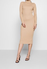Abito in maglia a coste beige con collo alto, maniche lunghe e silhouette aderente, che arriva a metà polpaccio, abbinato a sandali neri con tacco.