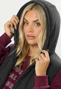 Svart huvtröja med dragkedja fram, som har en vadderad design. Modellen bär en bordeauxfärgad sweatshirt under, som visar vita textdetaljer.