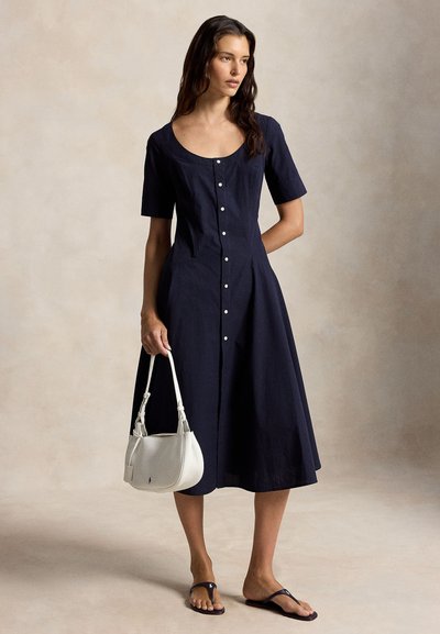 Γυναίκα που φοράει midi φόρεμα σε μπλε ναυτικό χρώμα με κουμπιά στο μπροστινό μέρος και κοντά μανίκια, κρατάει μικρή λευκή τσάντα χειρός και φοράει μαύρα σανδάλια, στέκεται μπροστά σε ουδέτερο φόντο.