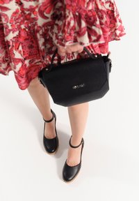 Borsa a tracolla in pelle nera con un design strutturato e un sottile dettaglio del logo. Scarpe nere con cinturini alla caviglia e una parte superiore testurizzata. Vestito floreale.