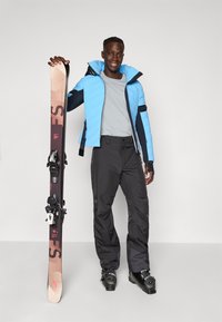 Homme souriant portant une veste d'hiver bleue, un pantalon de neige noir et des bottes de ski, tenant un ski en position verticale sur un fond clair.