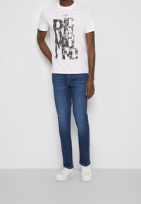 Witte katoenen T-shirt met een opvallend zwart grafisch ontwerp met de tekst "JOHN RICHMOND," gecombineerd met donkerblauwe slim-fit jeans en witte sneakers.