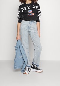 Black cropped long-sleeve shirt with white text, light blue high-waisted jeans, and black high-top sneakers. Denim jacket held in one hand.