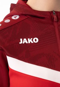 Rote Sportjacke aus strukturiertem Material mit Kapuze, weißem "JAKO"-Logo und einem horizontalen weißen Streifen über der Brust.
