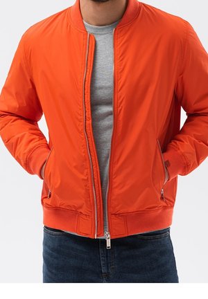 Man draagt een feloranje bomberjack over een lichtgrijs T-shirt en donkerblauwe spijkerbroek, handen in de jaszakken, ritssluiting half open.
