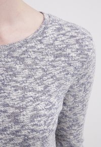 Gestrickter Pullover mit langen Ärmeln und Rundhalsausschnitt, der ein gesprenkeltes Muster in hellgrauen und marineblauen Farbtönen zeigt. Weiche Textur.