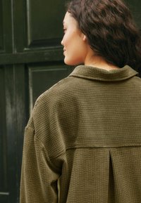 Camicetta verde oliva, con texture a coste e colletto, caratterizzata da un dettaglio a pieghe verticali sul retro e una vestibilità rilassata.