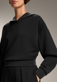 Felpa nera in cotone con cappuccio, vestibilità rilassata e maniche ampie. Tessuto liscio, dettagli delle cuciture minimi, indossata con pantaloni a vita alta.