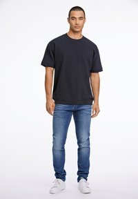 Felpa corta a maniche corte di colore blu scuro con scollatura rotonda, abbinata a jeans slim-fit di colore azzurro chiaro e sneakers bianche, su uno sfondo bianco.