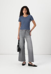 Blå randig kortärmad topp, grå vida jeans och svarta platta skor med ögla-accenter. Håller en liten svart handväska.