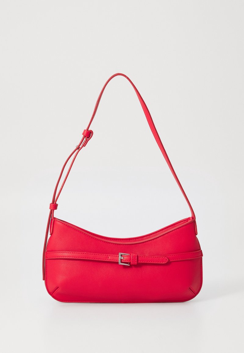 Sac à main rouge porté à l'épaule avec une lanière ajustable fine et une ceinture décorative avec une boucle argentée sur le devant, sur un fond uni.