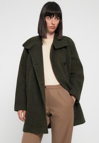 Olive green wool blend coat with a wide collar, double-breasted buttons, and a relaxed fit, paired with light beige top and brown pants.