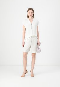Hvit, kortermet topp med glidelåsdetalj, kombinert med lysebeige shortser. Modellen holder en liten lavendelfarget håndveske og har på seg sko med klare stropper.