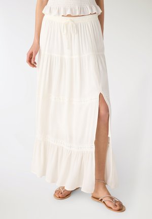 Donna che indossa una maxi gonna a balze bianca con spacco laterale, abbinata a una maglietta bianca con volant e sandali piatti color cuoio.