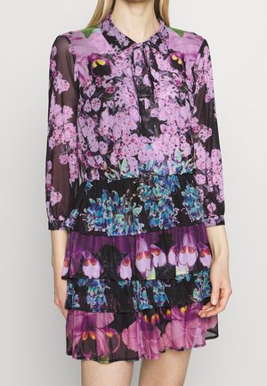 Femme portant une robe à manches longues imprimée florale avec une jupe à volants dans des nuances de violet, rose, bleu et noir, avec un col et un tissu transparent.