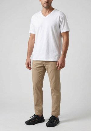 Mand iført hvid V-hals T-shirt, beige bukser og sorte lukkede sandaler, stående mod en lysegrå baggrund.