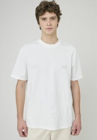 T-shirt blanc en coton avec un col rond, coupe décontractée, comportant un texte rose clair qui dit "DÉCONTRACTÉ" et "COURTS DE PADEL" sur le devant.