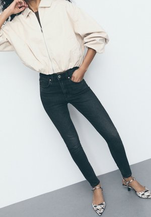 Persona que lleva una chaqueta beige con cremallera, vaqueros pitillo negros y tacones puntiagudos con patrón en negro y blanco, posando contra una pared blanca.