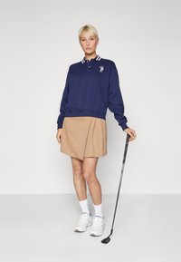 Tengerészkék pulóver csíkos gallérral és hímzett mintával, egy bézs redős szoknyával párosítva. Fehér tornacipők és egy golfütő teszi teljessé az öltözetet.