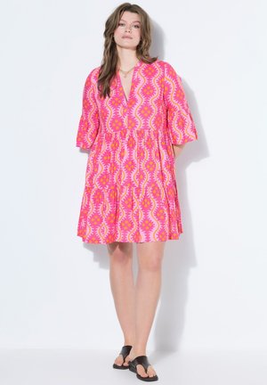 Kobieta stojąca, ubrana w różowo-pomarańczową sukienkę za kolano z rozkloszowanymi rękawami i czarne wsuwane sandały na białym tle.