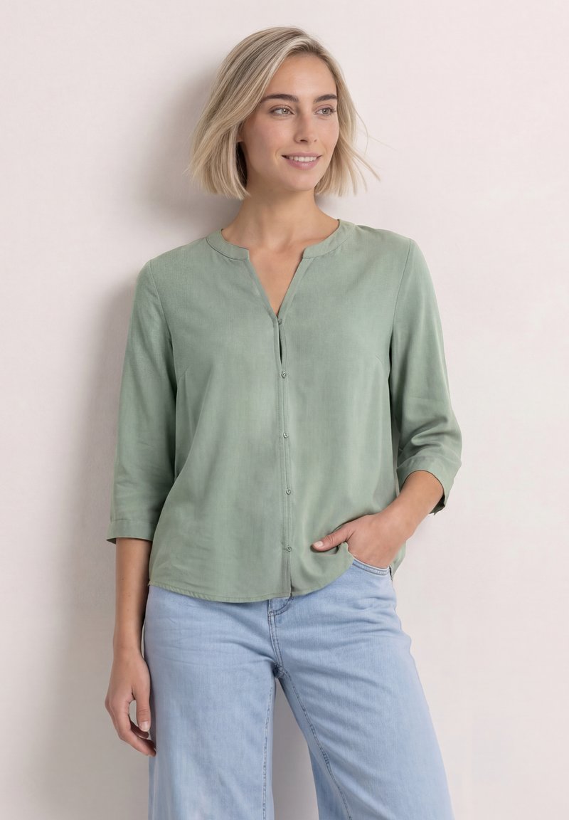 Femme aux cheveux blonds courts portant un chemisier vert clair boutonné et un jean bleu clair, debout avec une main dans la poche devant un fond uni.