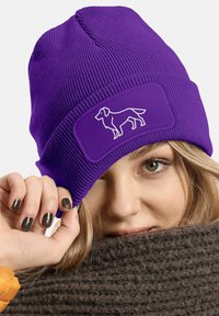Purple knitted beanie with a rectangular patch featuring a white dog silhouette. The wearer holds the brim, showcasing dark, glittery nails.