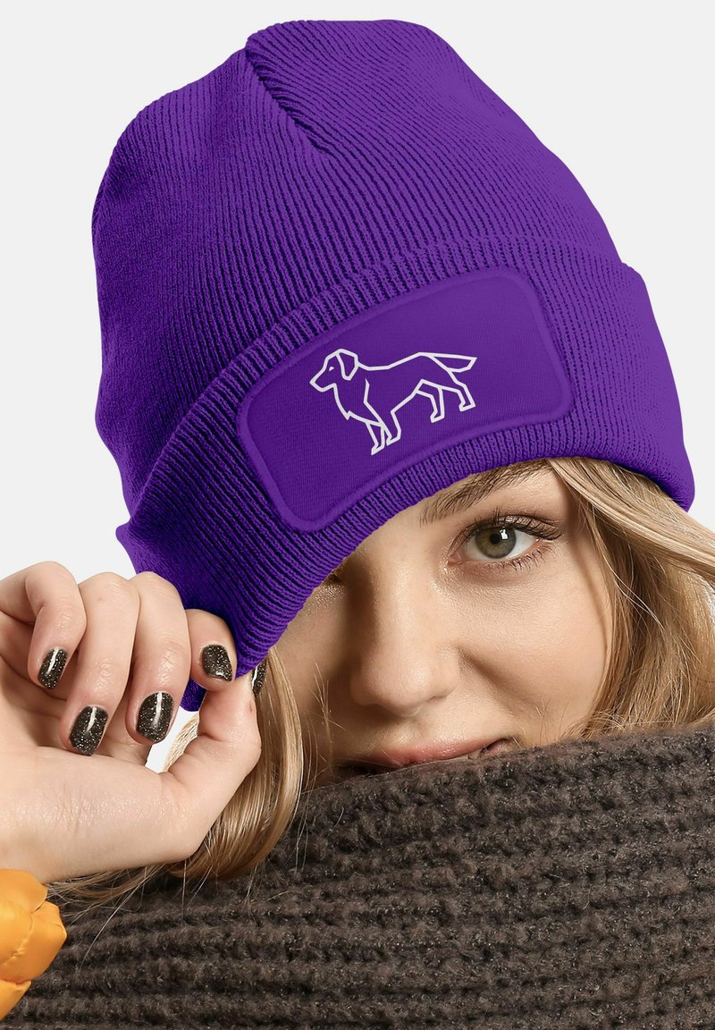 Purple knitted beanie with a rectangular patch featuring a white dog silhouette. The wearer holds the brim, showcasing dark, glittery nails.