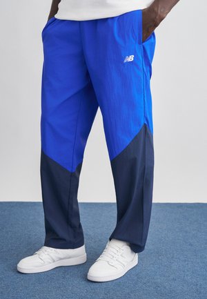 Person wearing blue and dark navy tapered pants with white New Balance sneakers, standing on blue carpet with hands in pockets.