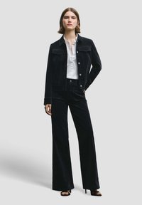 Giacca in velluto a coste nero con pantaloni abbinati a gamba larga, abbinati a una camicia bianca con bottoni. Texture liscia, vestibilità sartoriale e bottoni d'argento.