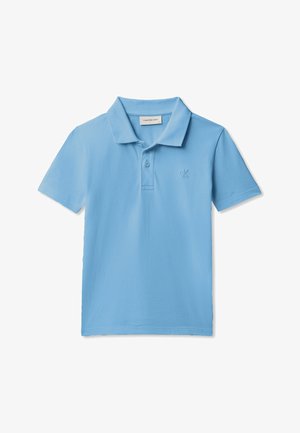 Junge mit lockigem Haar, der ein hellblaues Polohemd, graue Jeans, schwarze Schuhe trägt und eine schwarze Jacke um die Taille hält.