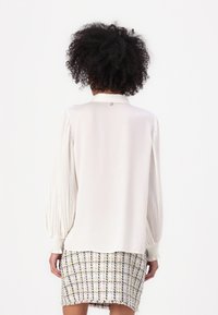 Blusa blanca con mangas largas plisadas y un cuello clásico. Se combina con una falda de tweed texturizada y multicolor.