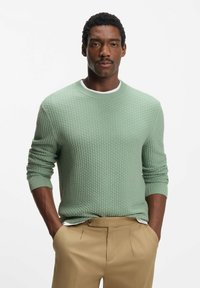 Vaaleanvihreä, kuvioitu neulepaita, jossa pyöreä pääntie ja resorireunaiset hihat, yhdistetty beigen värisiin housuihin. Kankaassa on hienovarainen kudottu kuvio.