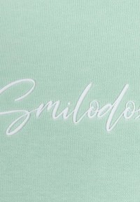 Il tessuto verde menta presenta una superficie liscia con lettere bianche in rilievo che compongono la parola "Smilodon". Il design è semplice e moderno.