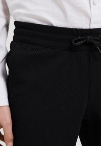Pantaloni jogging neri con texture a waffle, dotati di vita elasticizzata e chiusura con stringa. Abbinati a una camicia bianca con bottoni, mostrando la posizione delle mani.